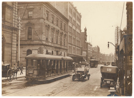 A sepia photograph of a city street lined with sandstone buildings and tram tracks running down the road. Two trams travel away from the viewer on the left track while a car travels towards the viewer on the right side.